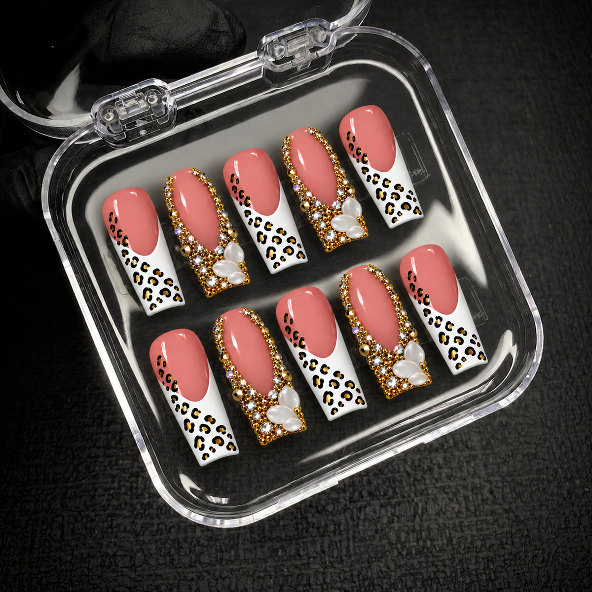 Open clear case holding ten coordinated blush pink leopard-print and jeweled nails on a dark textured background.
