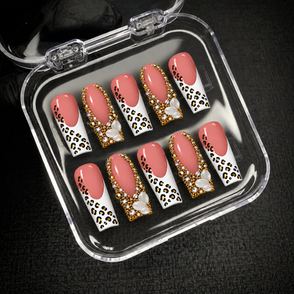 Open clear case holding ten coordinated blush pink leopard-print and jeweled nails on a dark textured background.