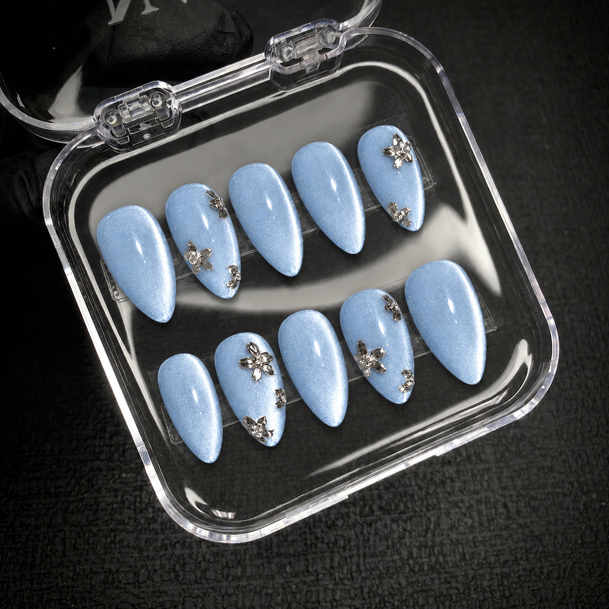 Open clear display case showing ten pale blue shimmer nails with silver-toned floral accents against a dark background.