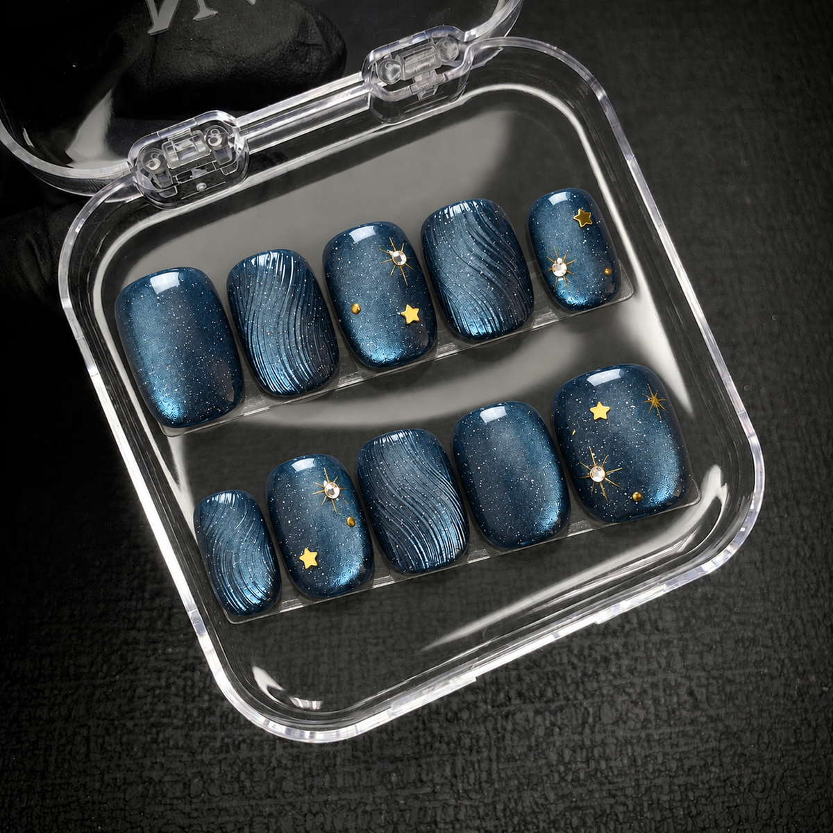 Ten navy-blue nails with curved textures and gold star accents displayed inside an open clear case.