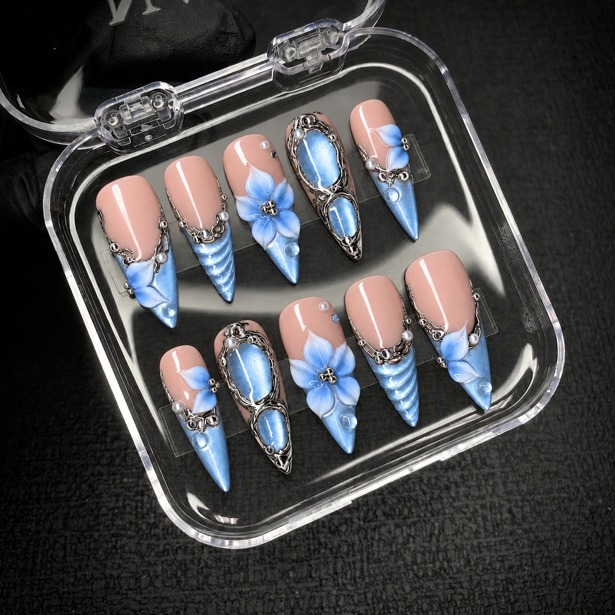 Open clear case on a dark background displaying ten pink-and-blue press-on nails with floral details and silver-tone ornament.