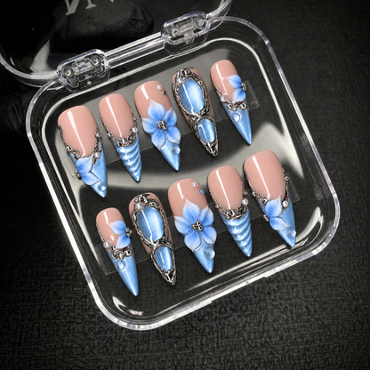 Open clear case on a dark background displaying ten pink-and-blue press-on nails with floral details and silver-tone ornament.
