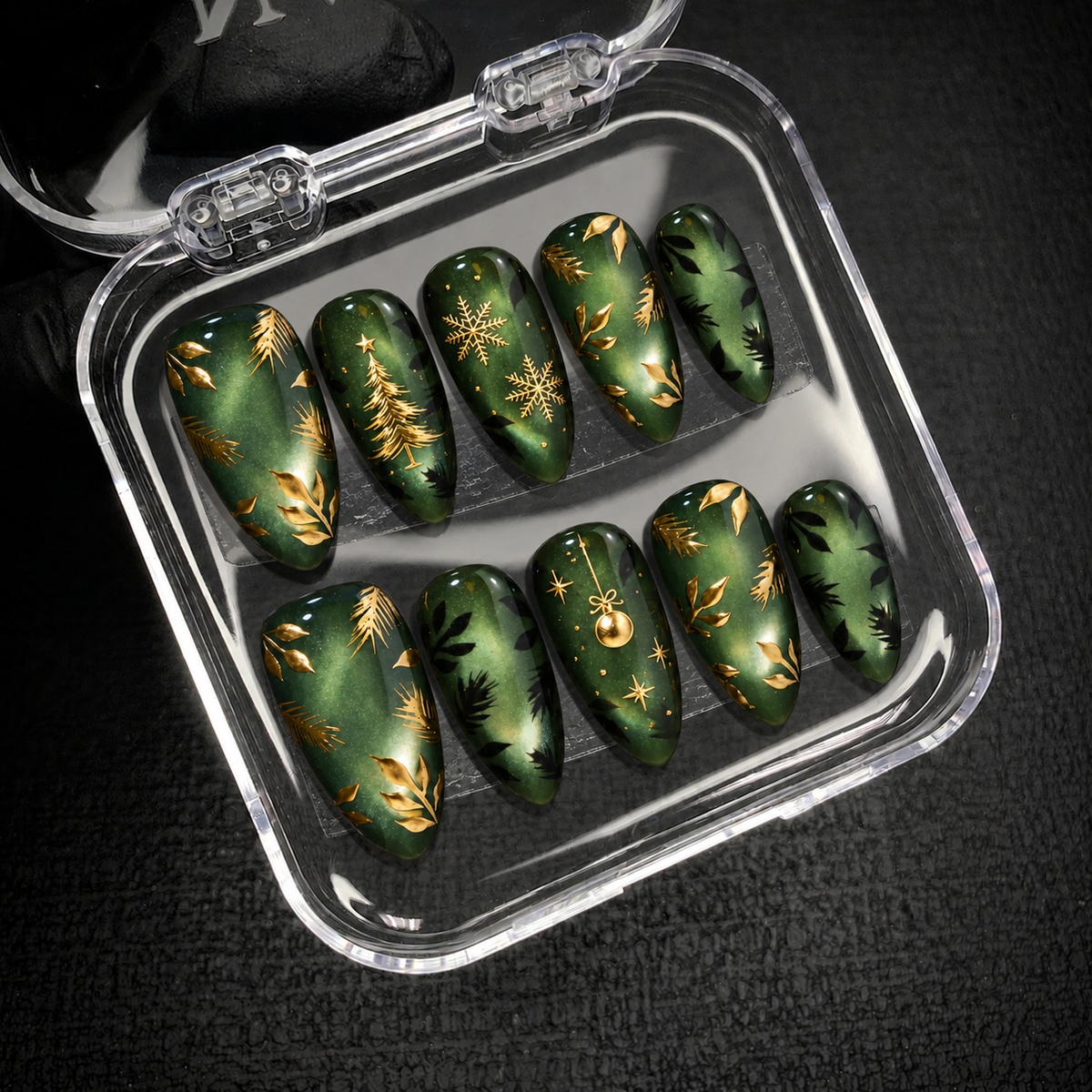 Open clear case holding ten green and gold Christmas nails in two rows on dark textured fabric.