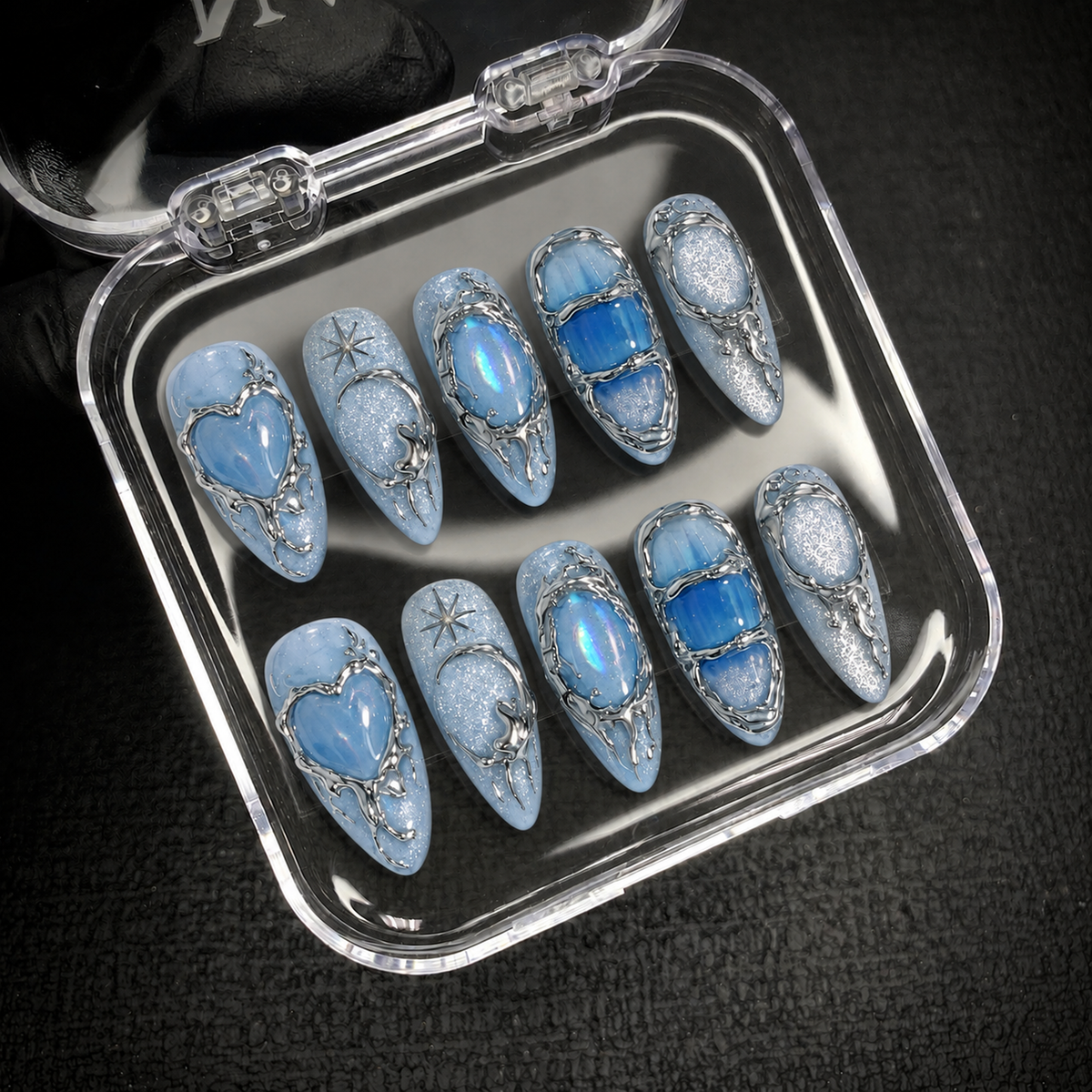Open clear case displaying ten coordinated icy blue nails with heart, crescent, oval, and blue panel designs.