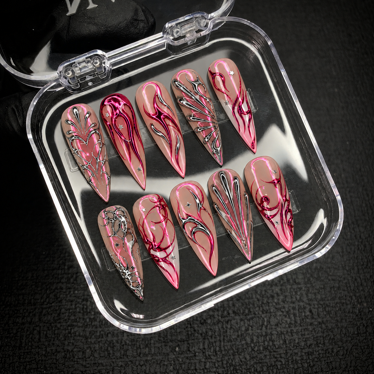 Open clear case holding ten pointed nails with pink curves, silver branching patterns, and star accents on a dark textured surface.