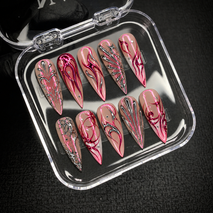 Open clear case holding ten pointed nails with pink curves, silver branching patterns, and star accents on a dark textured surface.
