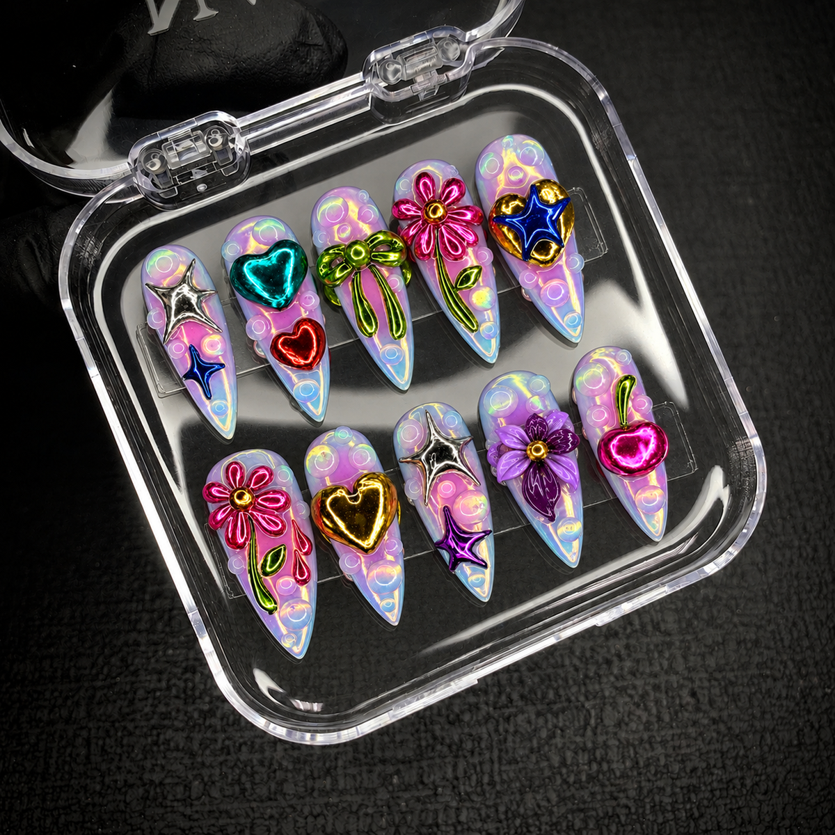 Open clear case on dark textured fabric holding ten coordinated nails with pastel bubble details and bright metallic floral, heart, star, bow, and cherry accents.