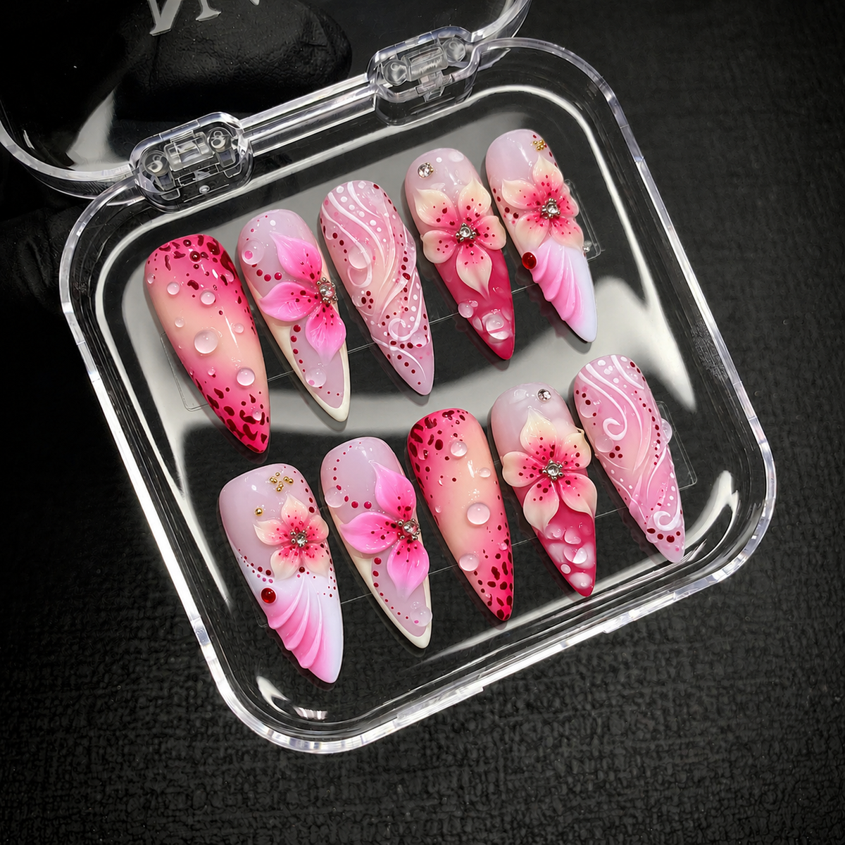 Open clear nail case on a black textured surface holding ten coordinated pink nails with raised flowers, white swirls, and droplet accents.