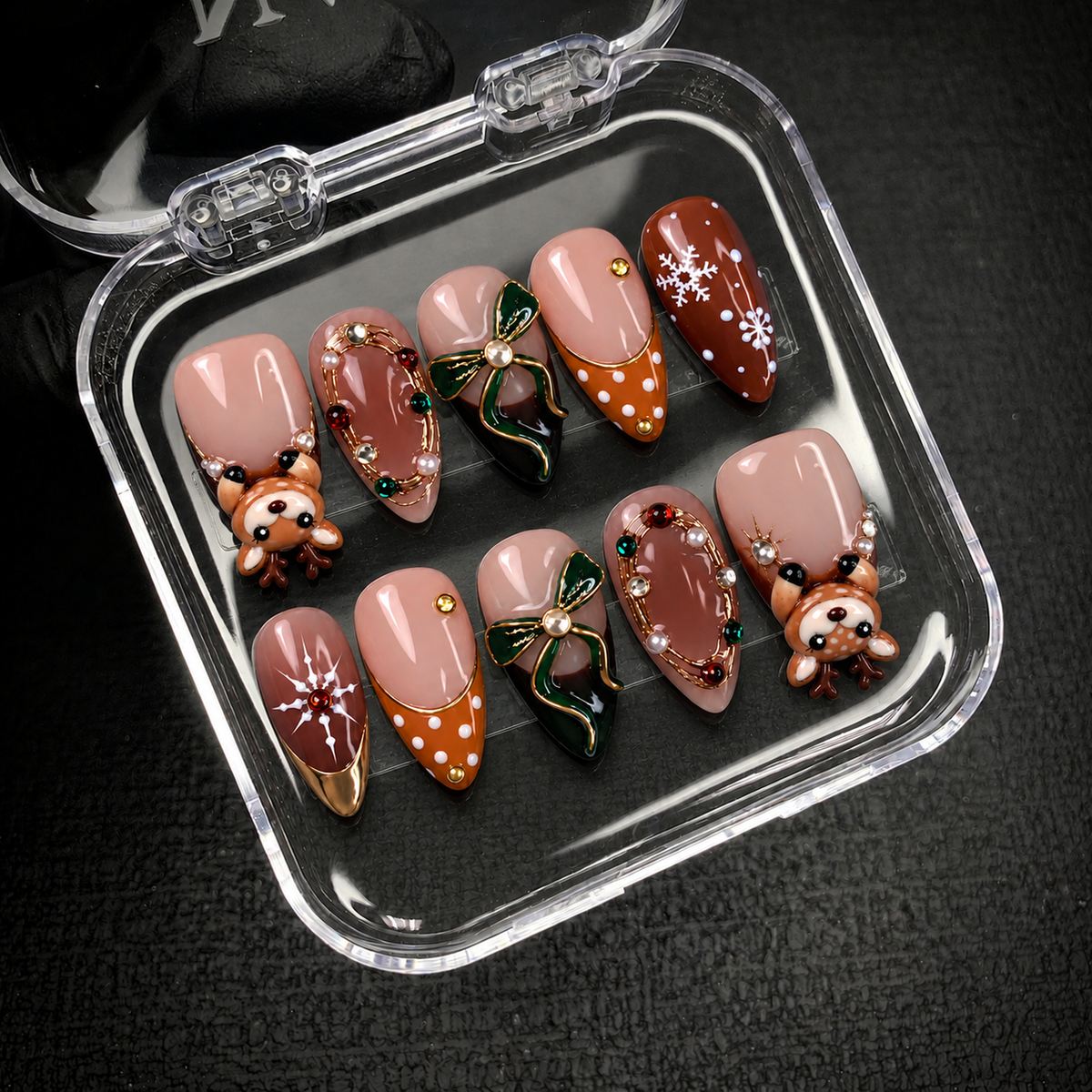 Open clear case on a dark textured surface holding ten coordinated nails with raised reindeer, green bows, and blush and brown Christmas accents.