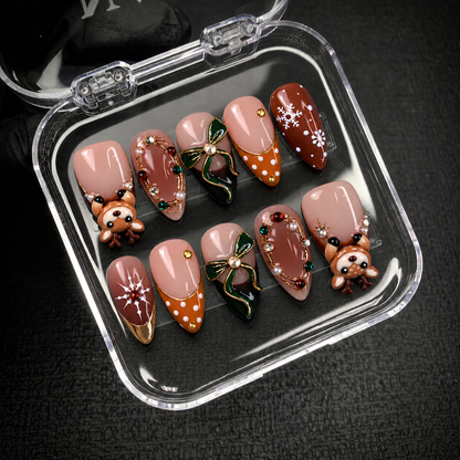 Open clear case on a dark textured surface holding ten coordinated nails with raised reindeer, green bows, and blush and brown Christmas accents.