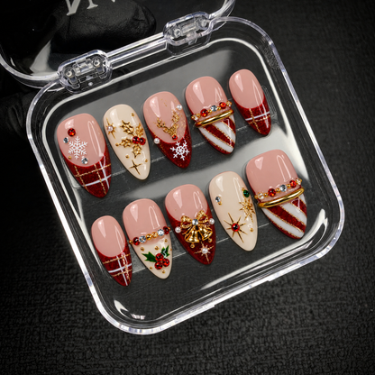 Open clear nail case on a dark textured surface holding ten coordinated blush, ivory, and ruby glitter Christmas nails with dimensional gold-tone accents.