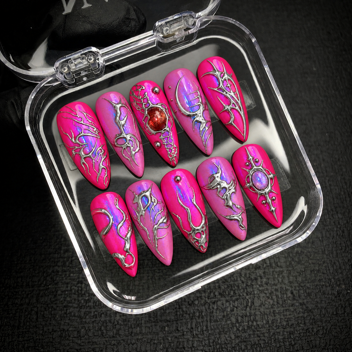 Open clear case holding ten coordinated pink nails with intricate silver-toned patterns and jewel-like accents on a dark textured surface.