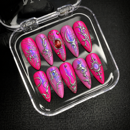 Open clear case holding ten coordinated pink nails with intricate silver-toned patterns and jewel-like accents on a dark textured surface.