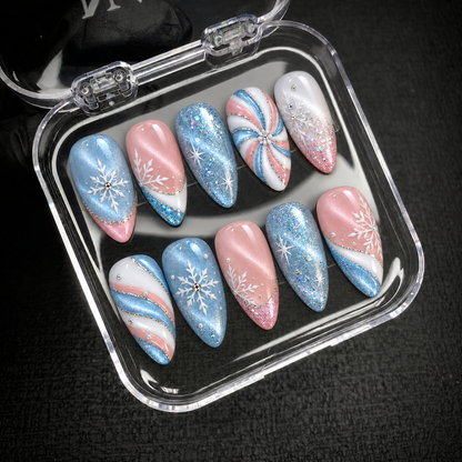 Open clear nail case on a dark textured surface holding ten blue, pink, and white nails decorated with glitter, snowflakes, and swirls.