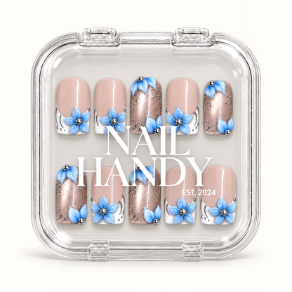 Clear branded case displaying ten Frosted Blue Petals nails in two rows, with blue flowers, glossy pink and white, and rose-toned shimmer accents.