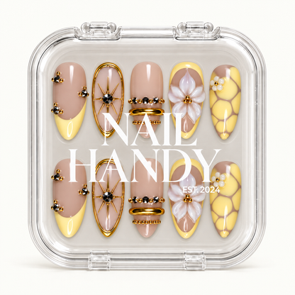 Closed clear case with white Nail Handy lettering, displaying ten yellow and nude nails with floral, starburst, and gold-tone bead accents.
