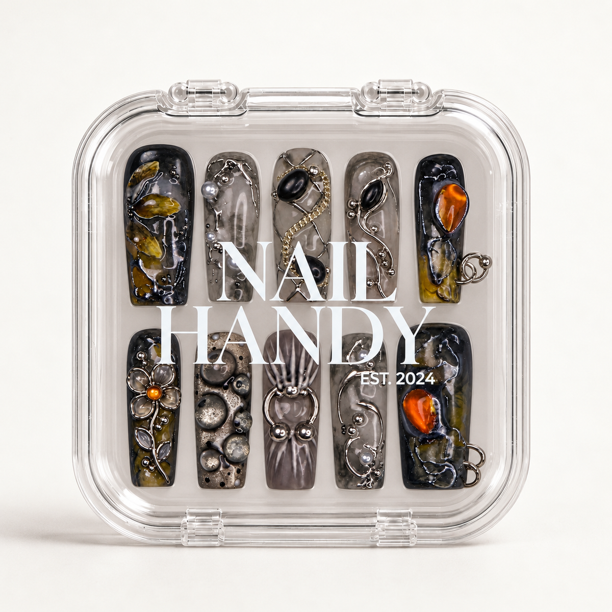 Closed clear case with white Nail Handy lettering, displaying ten coordinated nails with smoky backgrounds, orange accents, floral details, and metallic-looking embellishments.