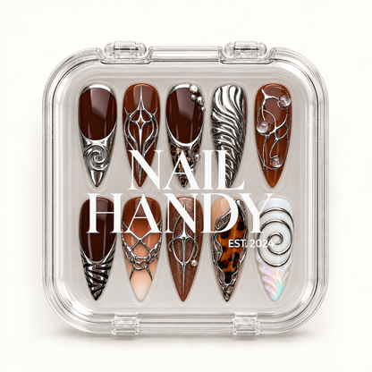Closed clear case displaying ten coordinated nails with brown bases, silver-toned sculptural accents, and a white spiral design beneath Nail Handy lettering.