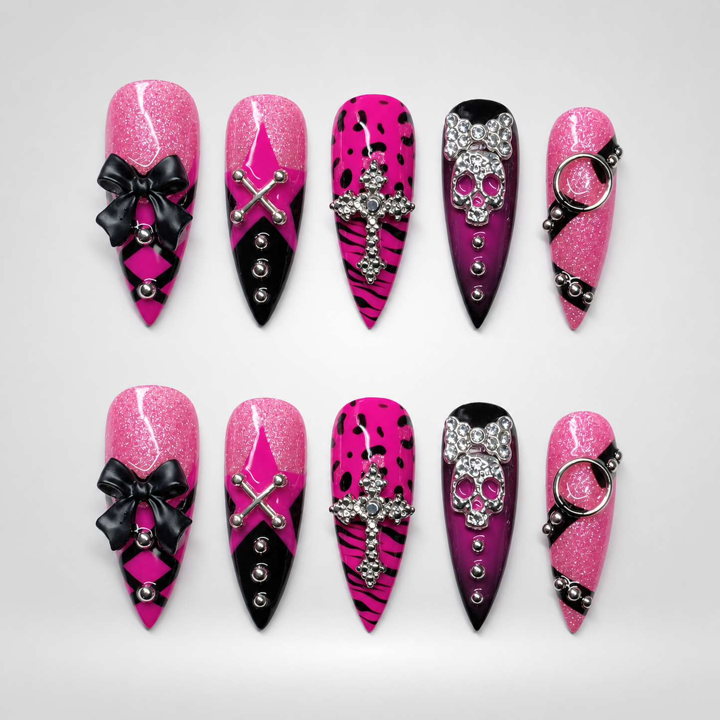 Ten coordinated pink glitter and black press-on nails displayed in two rows, featuring bows, silver-tone charms, crosses, spotted print, and skull-inspired details.