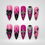 Ten coordinated pink glitter and black press-on nails displayed in two rows, featuring bows, silver-tone charms, crosses, spotted print, and skull-inspired details.