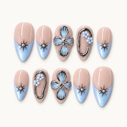 Ten blush-nude press-on nails arranged in two rows on a light background, with icy-blue tips, silver-tone starbursts, and blue floral accents.