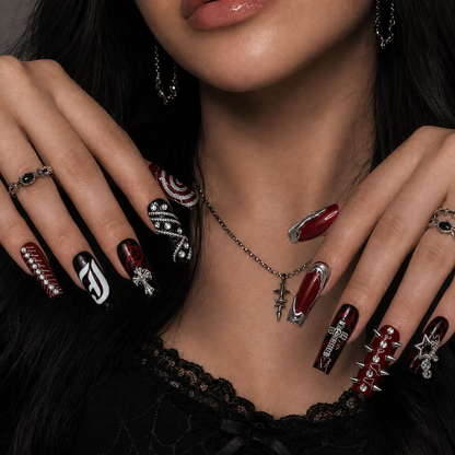 Hand model wearing Crimson Relic press-on nails with glossy burgundy and black flame artwork, chrome-tone crosses, chains, spikes, and crystal details.