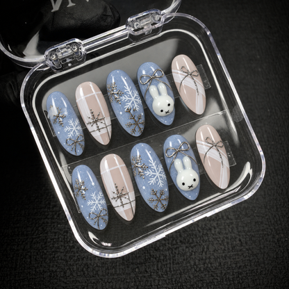 Open clear case displaying ten Snowbound Bunny nails in blue and nude with snowflakes, bows, plaid lines, and bunny motifs.