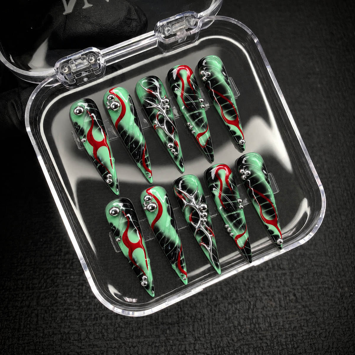 Open clear case holding ten mint green, black, and red pointed nails with silver accents, set diagonally on a dark textured surface.