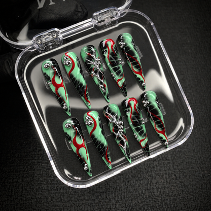 Open clear case holding ten mint green, black, and red pointed nails with silver accents, set diagonally on a dark textured surface.