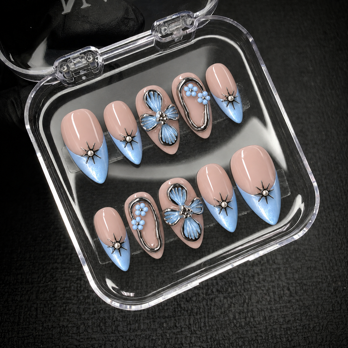Open clear nail case displaying ten blush-nude and icy-blue nails with silver-tone starbursts and blue flower accents against a dark background.