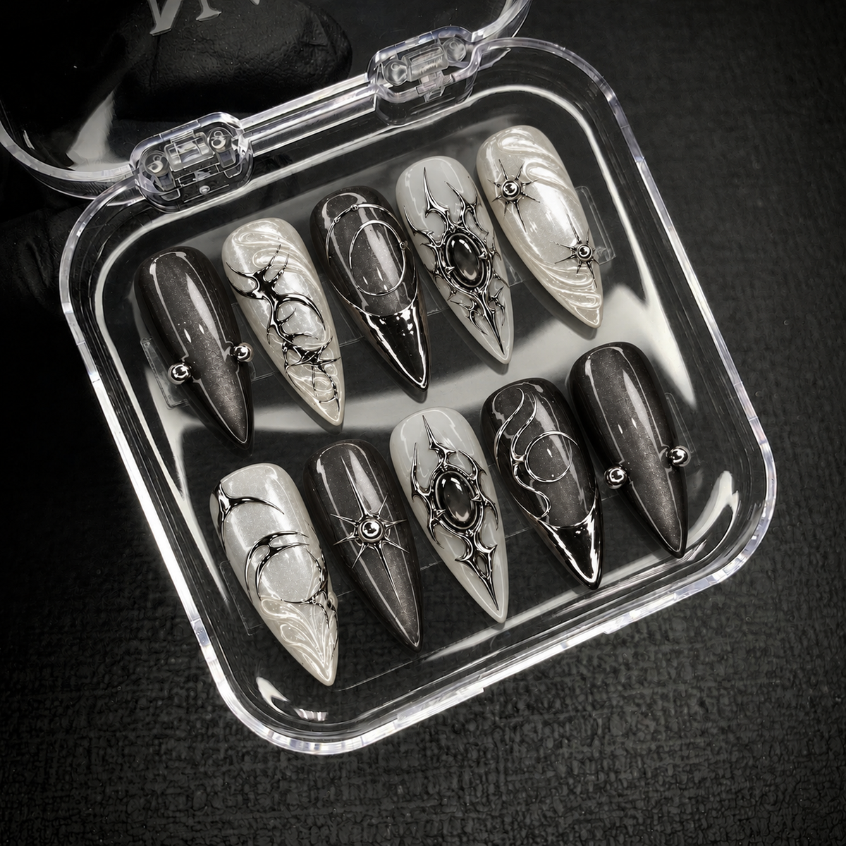 Open clear nail case on dark textured fabric displaying ten charcoal and pearly nails with silver-tone ornament, starbursts, and dark oval accents.