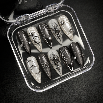 Open clear nail case on dark textured fabric displaying ten charcoal and pearly nails with silver-tone ornament, starbursts, and dark oval accents.