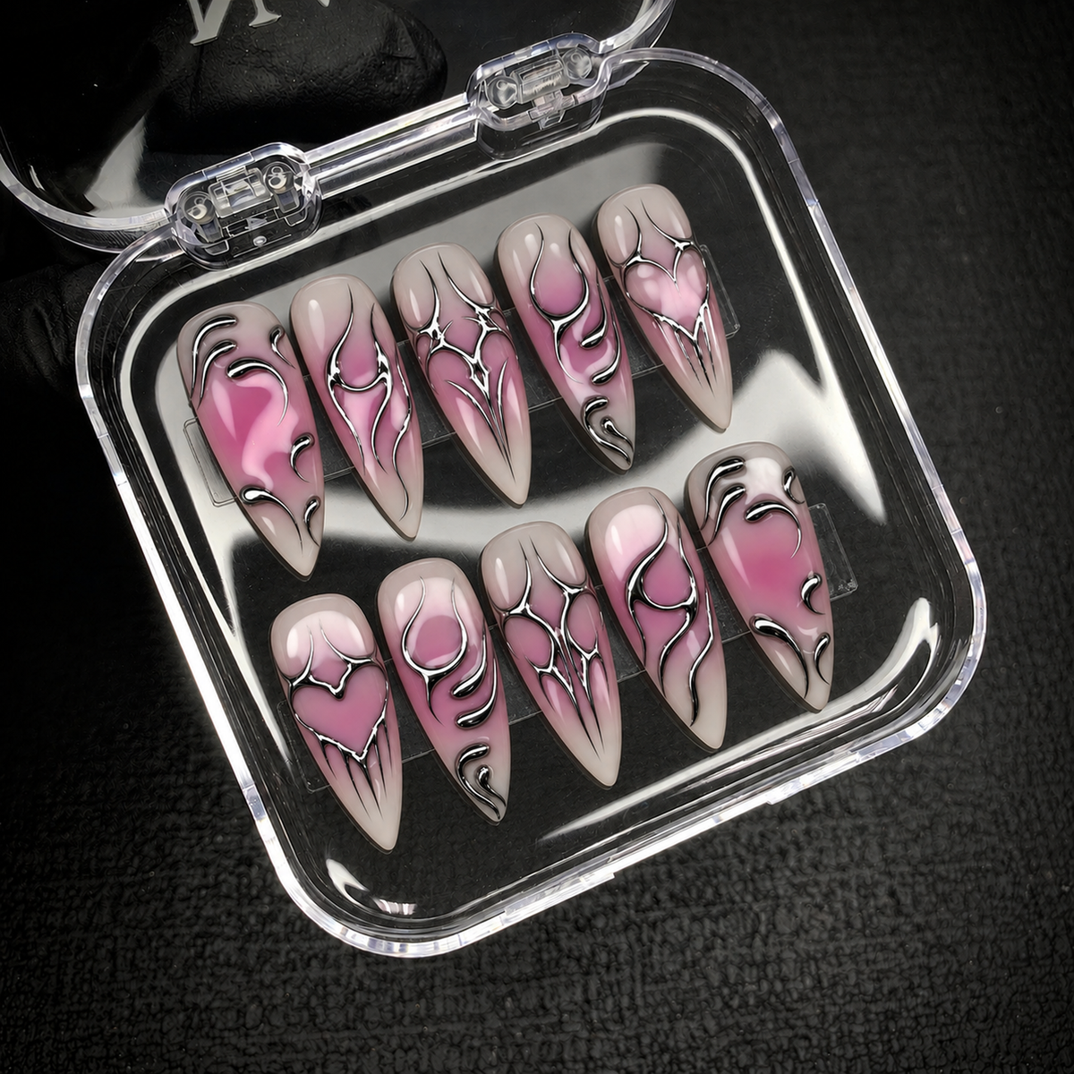 Open clear nail case on dark textured fabric holding ten pink, mauve, and greige nails with silver chrome-look hearts and curved accents.