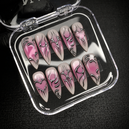 Open clear nail case on dark textured fabric holding ten pink, mauve, and greige nails with silver chrome-look hearts and curved accents.